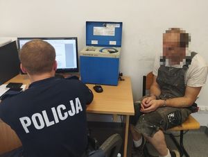 na zdjęciu widać policjanta siedzącego przy biurku, który pisze cos na komputerze. Obok policjanta siedzi mężczyzna z założonymi kajdankami.