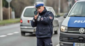 na zdjęciu widać policjanta ubranego w niebieska kurtkę z szarym napisem Policja. Policjant stoi na dworze w ręku trzyma miernik prędkości. Za policjantem widać drogę po lewej stronie przejeżdżający samochód natomiast po prawej radiowóz.