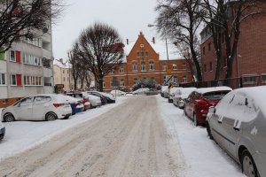 Na zdjęciu widać zaśnieżona drogę. Obok jezdni znajduje się parking na którym są zaparkowane pojazdy. W środkowej części zdjęcia w oddali widać budynek z cegły.