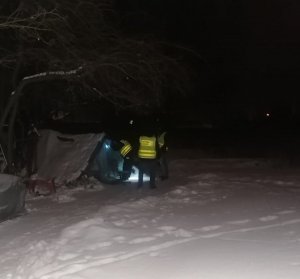 na zdjęciu widać jak funkcjonariusze w porze nocnej sprawdzają opuszczone miejsce, szałas. Funkcjonariusze świecą latarką. Dookoła leży duża warstwa śniegu.