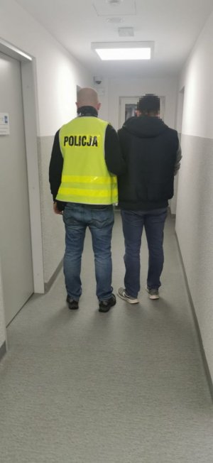 Na zdjęciu widać policjanta ubranego w kamizelkę żółtą z napisem policja oraz osobę zatrzymaną. Mężczyźni stoją tyłem. Osoba zatrzymana na rękach ma zapięte kajdanki. Stoją na korytarzu.