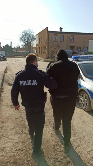 Na zdjęciu widać dwie osoby. Jedna z nich to funkcjonariusz policji natomiast druga jest to osoba zatrzymana. Osoba którą prowadzi umundurowany funkcjonariusz policji jest ubrana w czarna bluzę i czarne spodnie