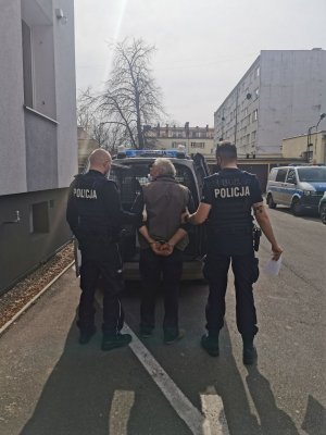 Na zdjęciu widać dwóch umundurowanych policjantów . Pomiędzy nimi stoi mężczyzna który ma założone kajdanki. W tle widać radiowóz policyjny oznakowany.