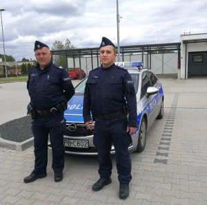 Na zdjęciu widać dwóch policjantów w umundurowaniu. Za funkcjonariuszami znajduje się oznakowany radiowóz policyjny.